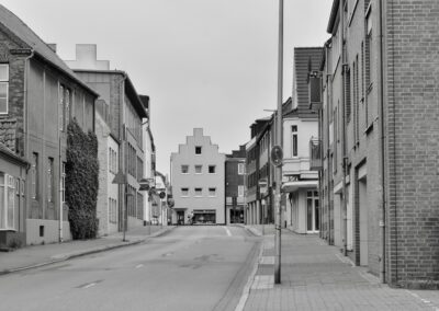 Die Schwarz-Weiß-Fotografie zeigt eine asphaltierte Straße, die im Hintergrund auf eine Querstraße trifft und endet. Die Straße ist auf beiden Seiten von Fußweg und Häusern umrahmt.