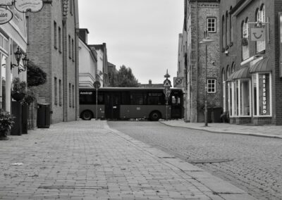 Die Schwarz-Weiß-Fotografie zeigt eine gepflasterte Straße, die sich von dem*der Betrachter*In wegbewegt. Sie wird von Fußweg auf beiden Seiten sowie Häusern umrahmt. Hinten im Bild kreuzt ein Linienbus die Straße. Vorne auf der linken Seite ist eine Stolperschwelle in den Fußweg eingelassen.